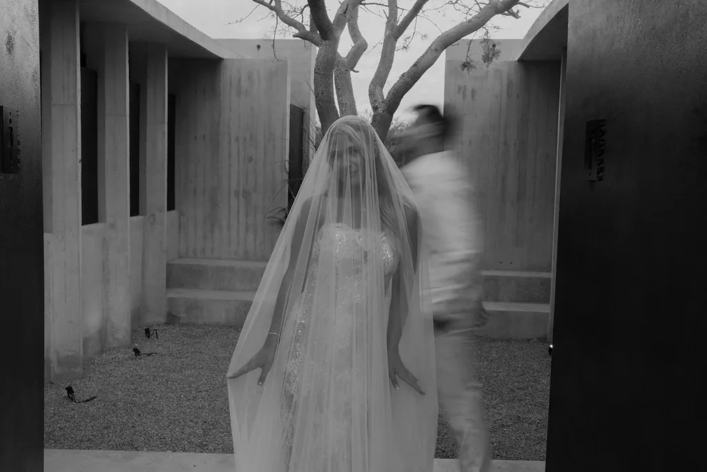 Bailey stands still in her veil while Matthew passes behind her, in black and white